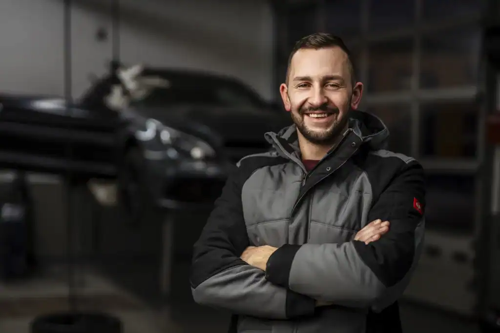 Martin Schwarz, Inhaber der SCHWARZ.KG Unfallinstandsetzung, lächelnd mit verschränkten Armen in einer Werkstatt vor einem unscharfen Fahrzeug.